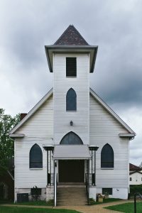A white wooden church building with a steep gabled roof and a small steeple on top.
