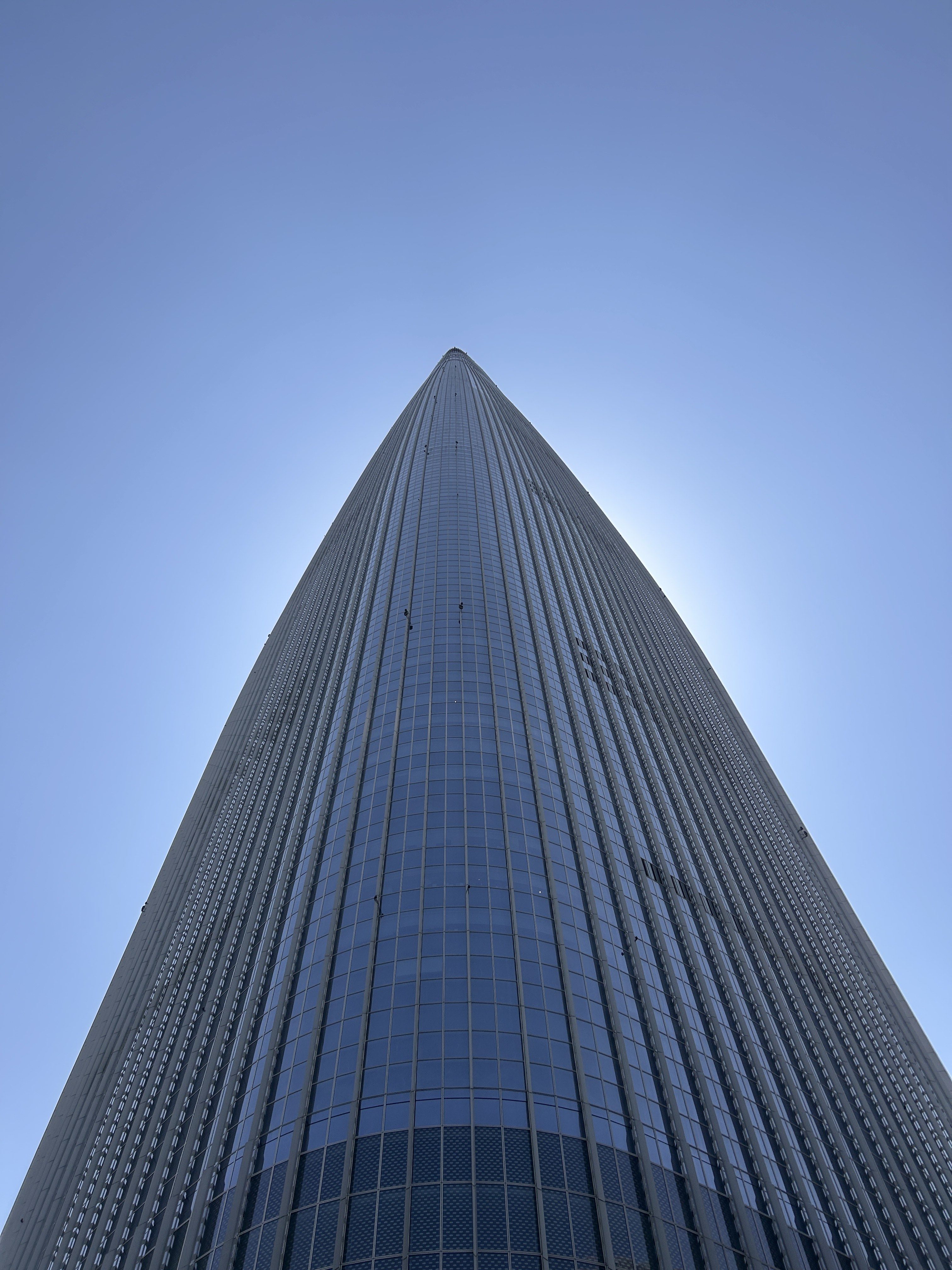 A tall, modern skyscraper viewed from the ground looking up, with a sharp, pointed top and a facade made chiefly of glass panels reflecting the clear blue sky.