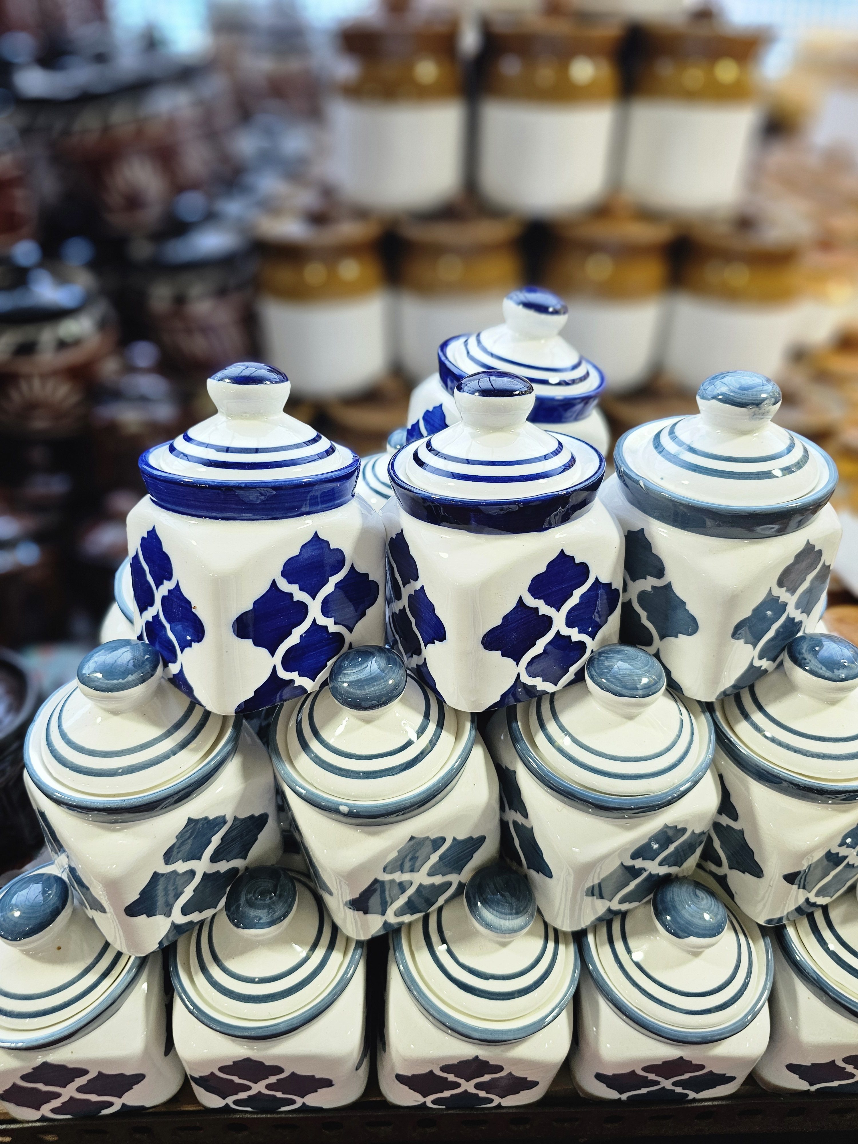 The image shows a collection of ceramic jars with lids, stacked together. The jars are mostly white with blue patterns, while some in the background are white with brown tops. Captured from Thrissur, Kerala.