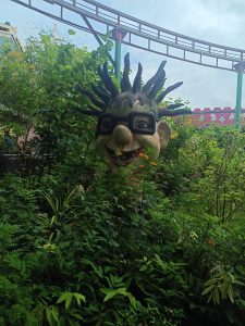 A large, whimsical sculpture of a cartoonish face with spiky hair and oversized glasses is partially obscured by lush green foliage and colorful flowers.