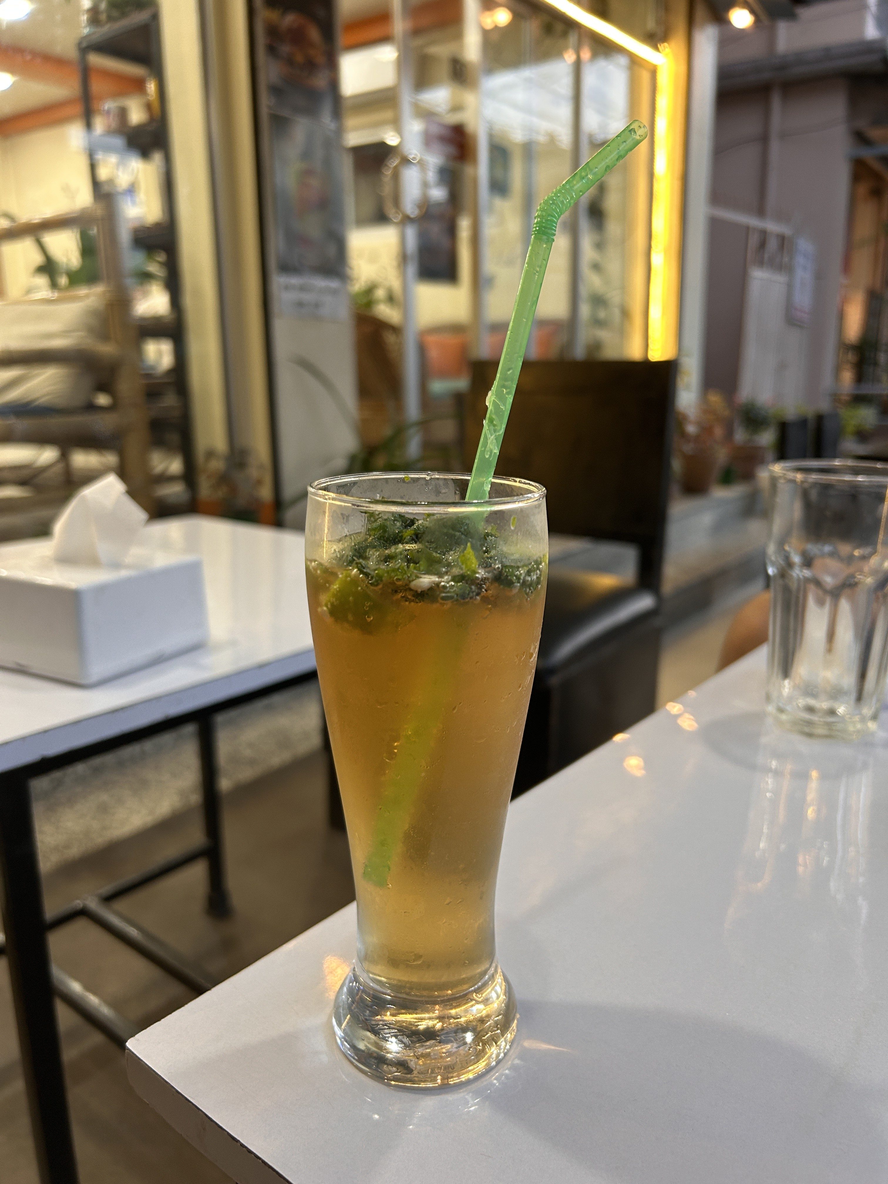A tall glass filled with a light brown beverage garnished with mint leaves and a green straw, placed on a white table.