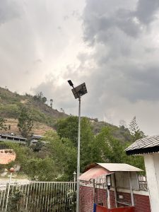 A tall solar panel mounted on a pole stands prominently against a backdrop of cloudy skies and a hilly landscape.
