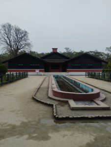 A view of a building with a dark wooden exterior and a thatched roof, situated in a landscaped area. In front of the building, there is a long, narrow water feature with a concrete border and several evenly spaced columns. 