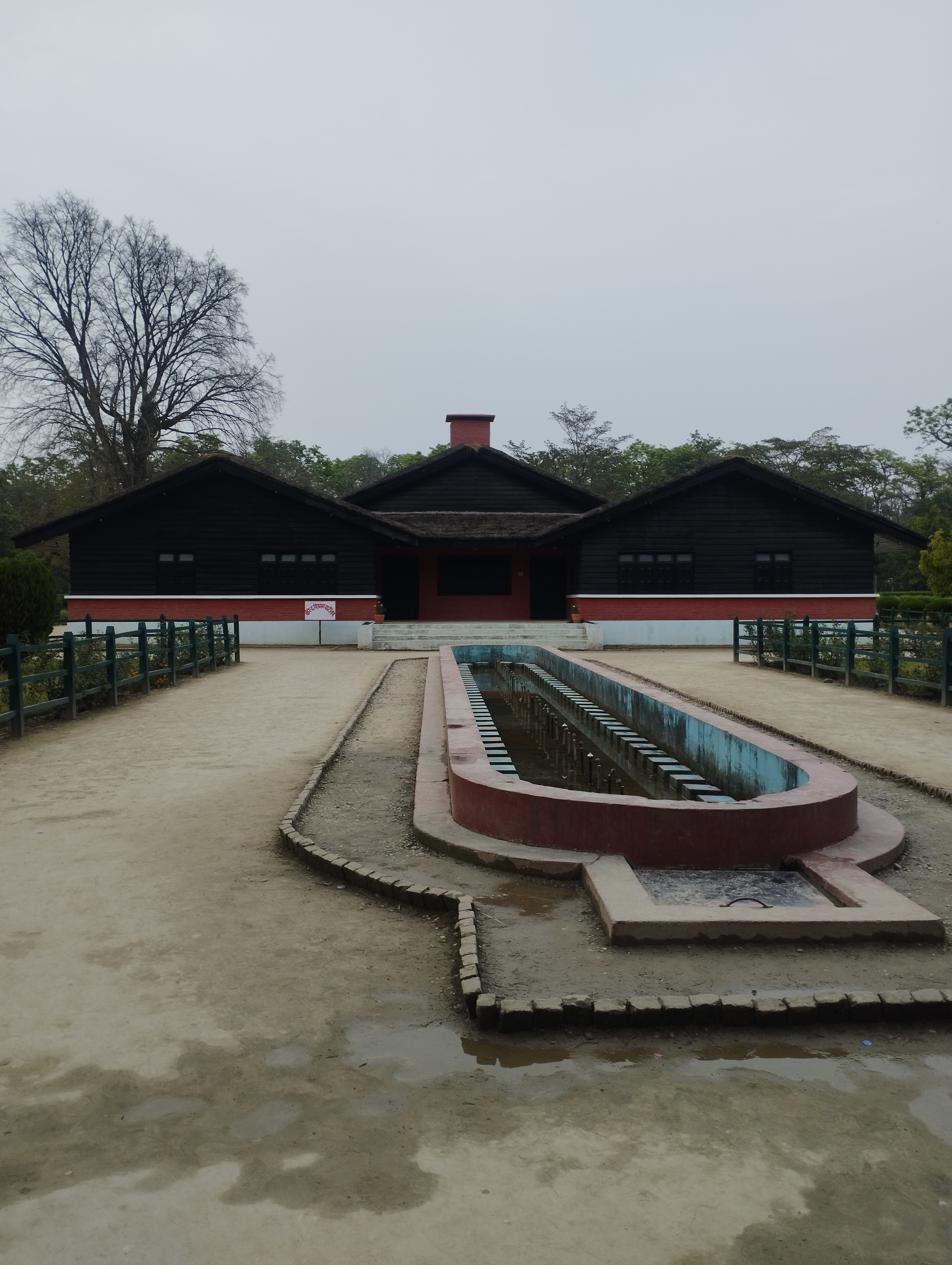 A view of a building with a dark wooden exterior and a thatched roof, situated in a landscaped area. In front of the building, there is a long, narrow water feature with a concrete border and several evenly spaced columns.
