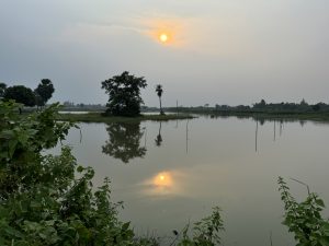 A tranquil landscape at sunset, featuring a calm body of water that reflects the darkening sky and the sun low on the horizon.
