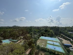 A panoramic view of a lush landscape under a bright blue sky dotted with clouds. In the foreground, there are several tennis courts with green and blue surfaces, surrounded by trees and vegetation.

