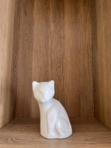 A white ceramic cat figurine sits on a wooden shelf with a natural wood grain texture.