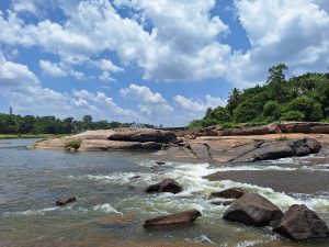 The image shows a river flowing over rocks under a partly cloudy sky. The water is clear, and the rocks are various sizes, with lush greenery visible on the riverbanks in the background.
