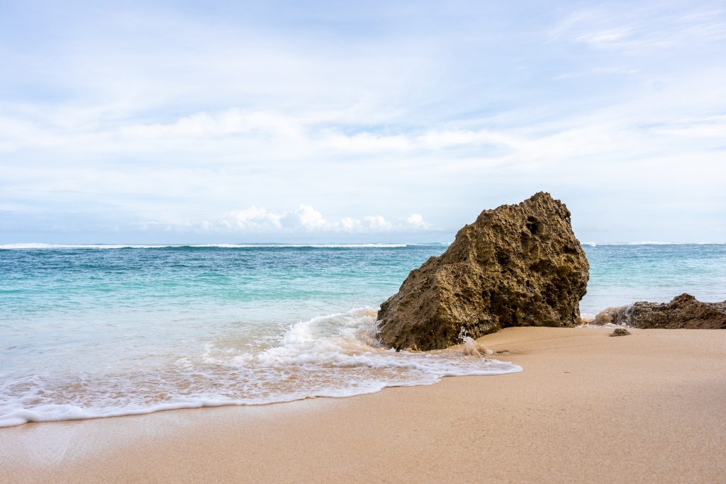 A rocky outcrop sits on a sandy beach, with gentle waves lapping at its base. The ocean stretches out in the background, displaying varying shades of blue and turquoise under a light cloudy sky.