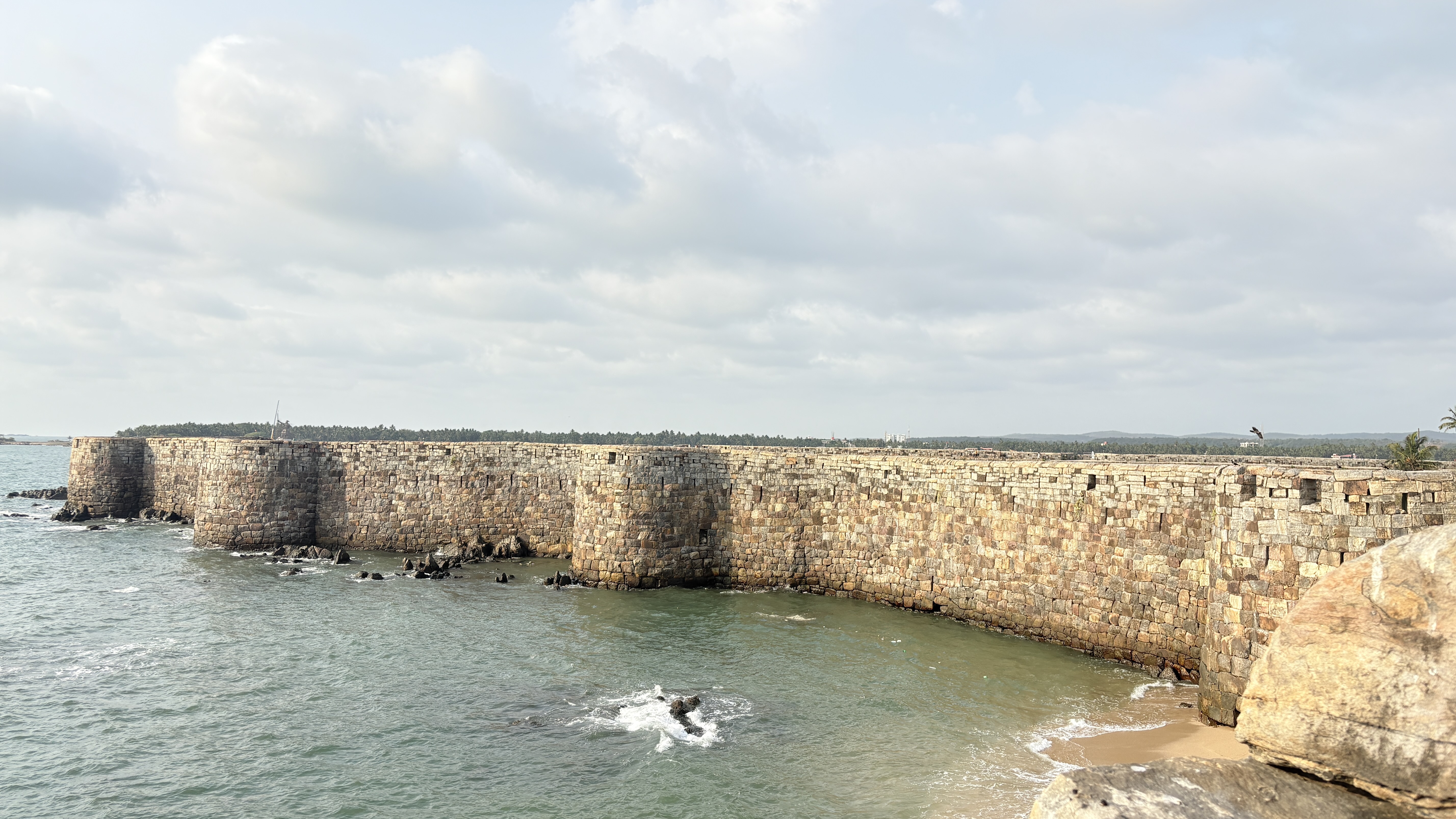 A long, curved stone wall stretches along the coastline, partially submerged in the shallow water. The wall features large, weathered stones and has several openings.