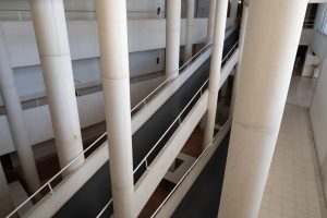 An interior view of a modern building featuring multiple levels connected by a long, narrow walkway. The space is supported by tall, white cylindrical columns, and there is a minimalistic design with smooth surfaces and a neutral color palette.