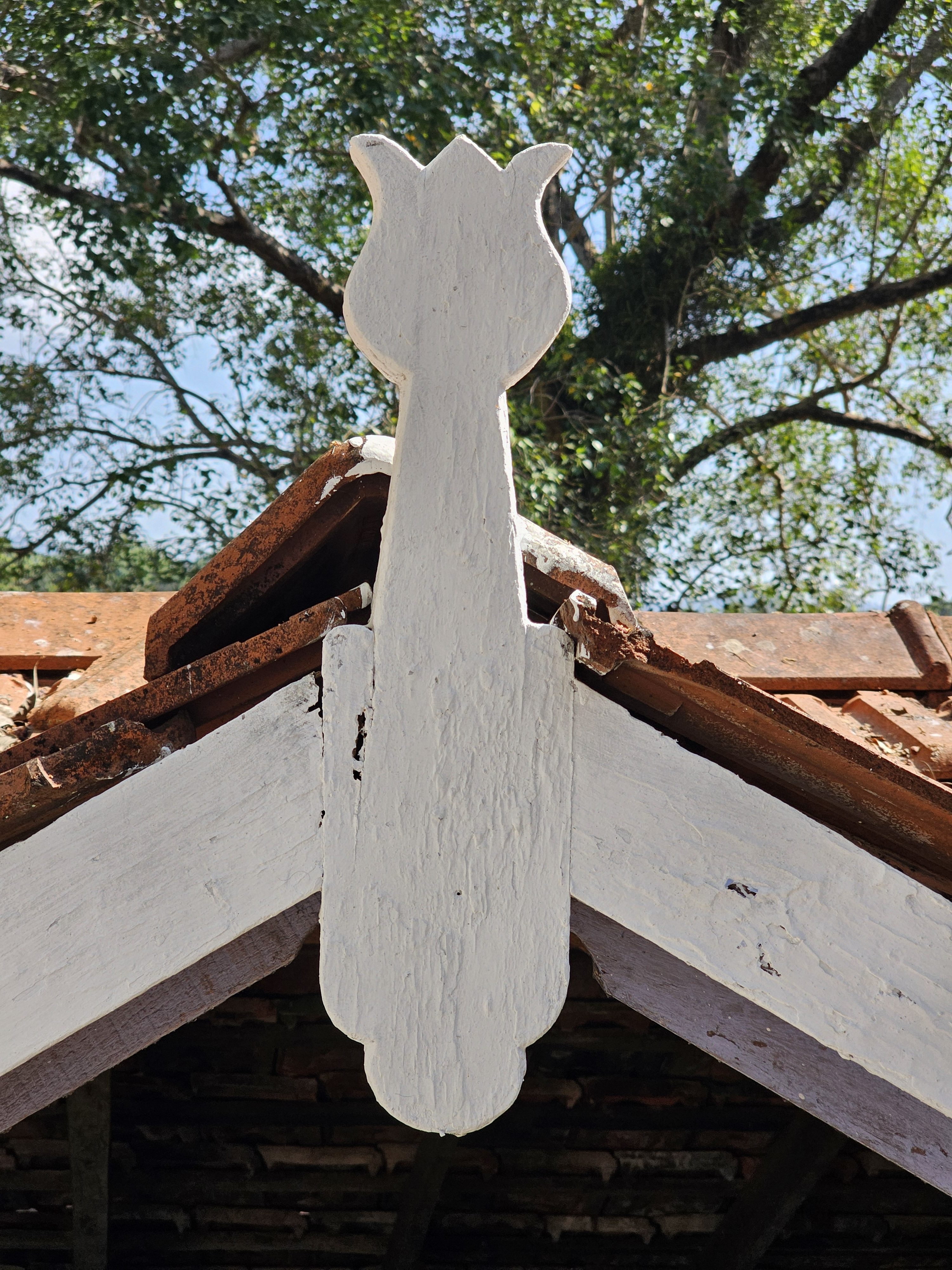 A close-up of a white, tulip-shaped wooden carving attached to the apex of a tiled roof, part of a traditional building near Thirumandhamkunnu Temple (Angadippuram, Malappuram, Kerala). The carving is positioned against a backdrop of green foliage and a partially visible blue sky, highlighting the architectural detail.