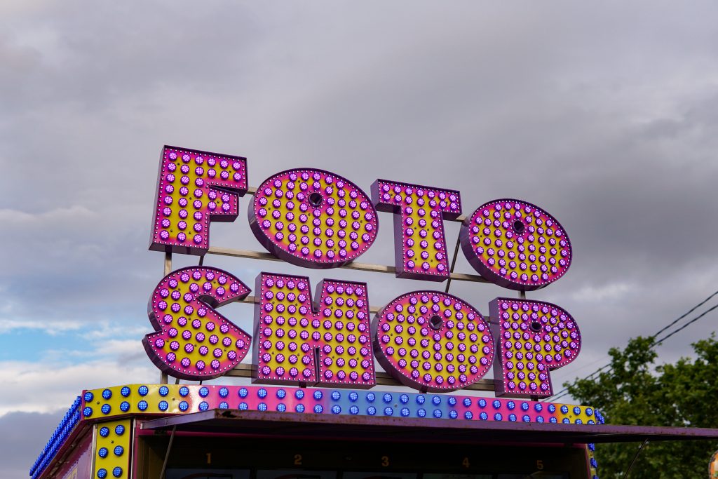 A brightly lit sign reading “FOTO SHOP” in large, bold letters adorned with colorful, flashing lights. The sign is mounted on a structure against a cloudy sky