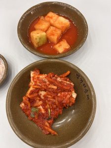 A close-up view of two small, round ceramic bowls on a light-colored table. One bowl contains cubes of pickled melon in a red sauce, while the other holds a piece of kimchi topped with shredded vegetables. The kimchi is rich in color, with hints of green from the added ingredients.