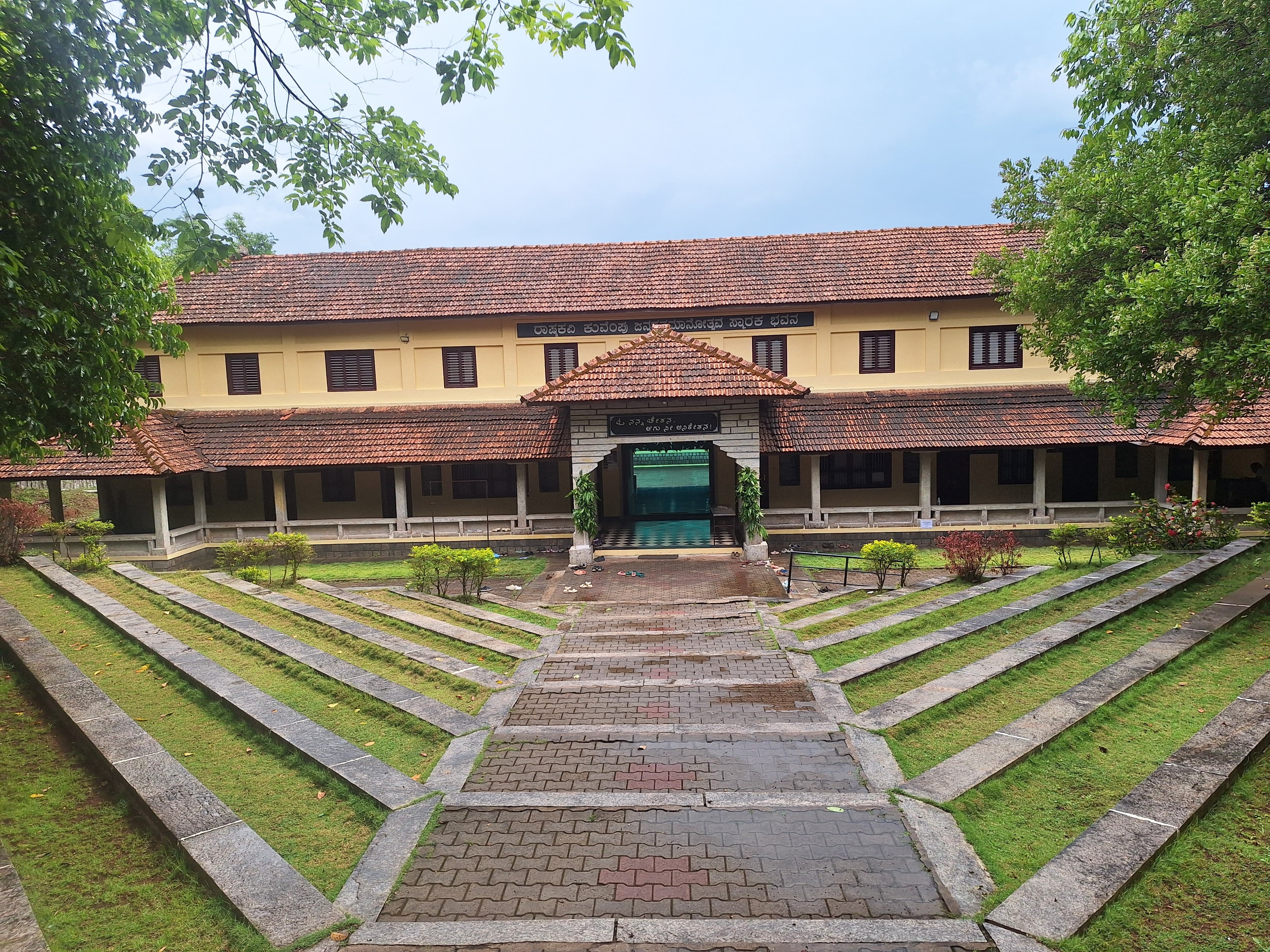 The image shows the Kuvempu Memorial Trust Ranga Mandira, a memorial hall dedicated to the Kannada writer and poet Kuvempu (Kuppalli Venkatappa Puttappa). The building features a traditional South Indian architectural style with a tiled roof and is surrounded by a well-maintained garden with geometric patterns.