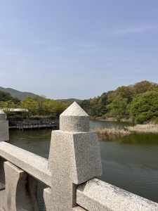 A close-up view of a granite railing with a decorative pointed pillar, overlooking a calm river. The background features lush greenery and wooded hills under a clear sky.