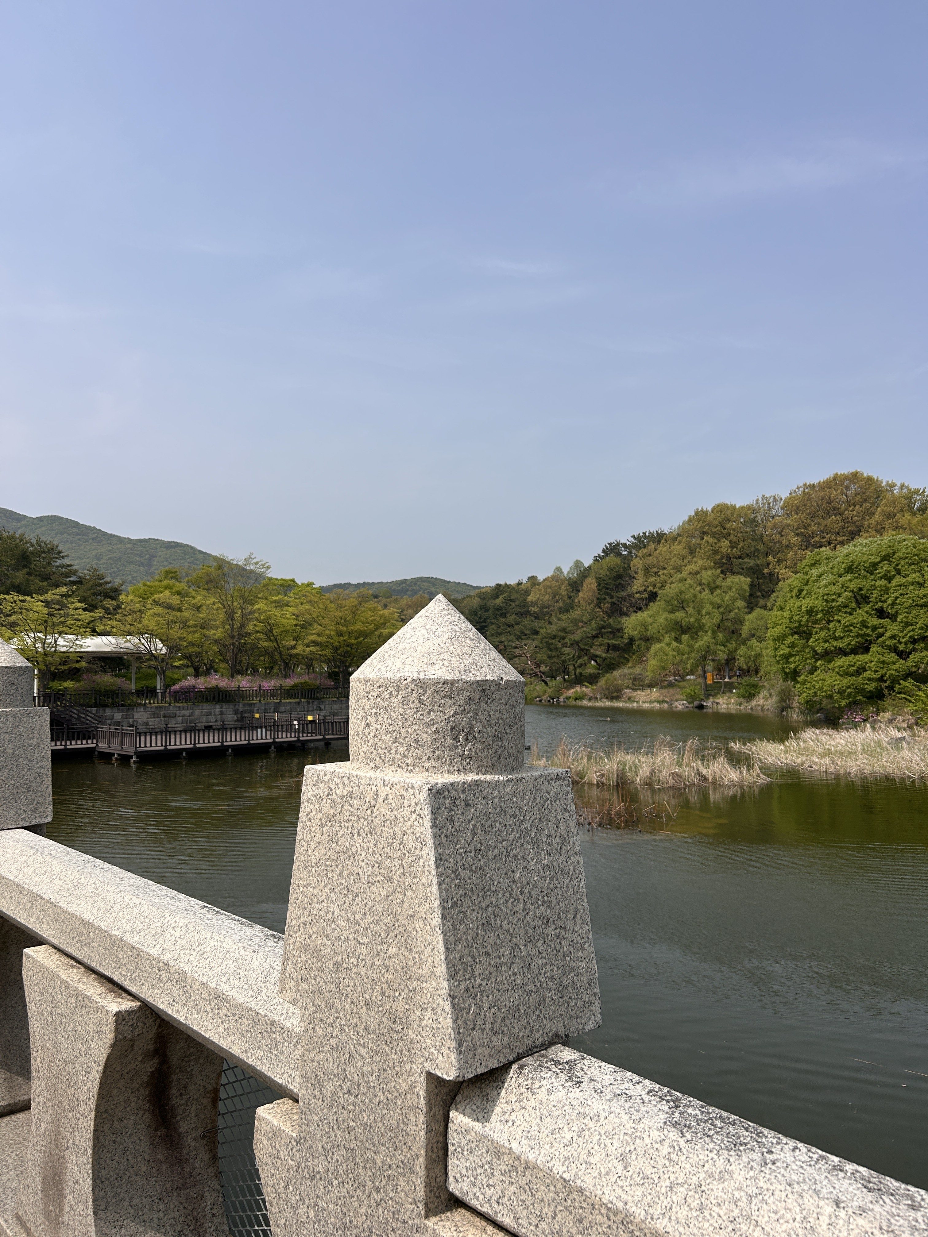 A close-up view of a granite railing with a decorative pointed pillar, overlooking a calm river. The background features lush greenery and wooded hills under a clear sky.
