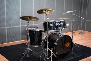 A black drum set is positioned on a black mat in a well-lit room with a wooden floor. The set includes a bass drum, snare drum, and two toms, accompanied by two cymbals mounted on stands.