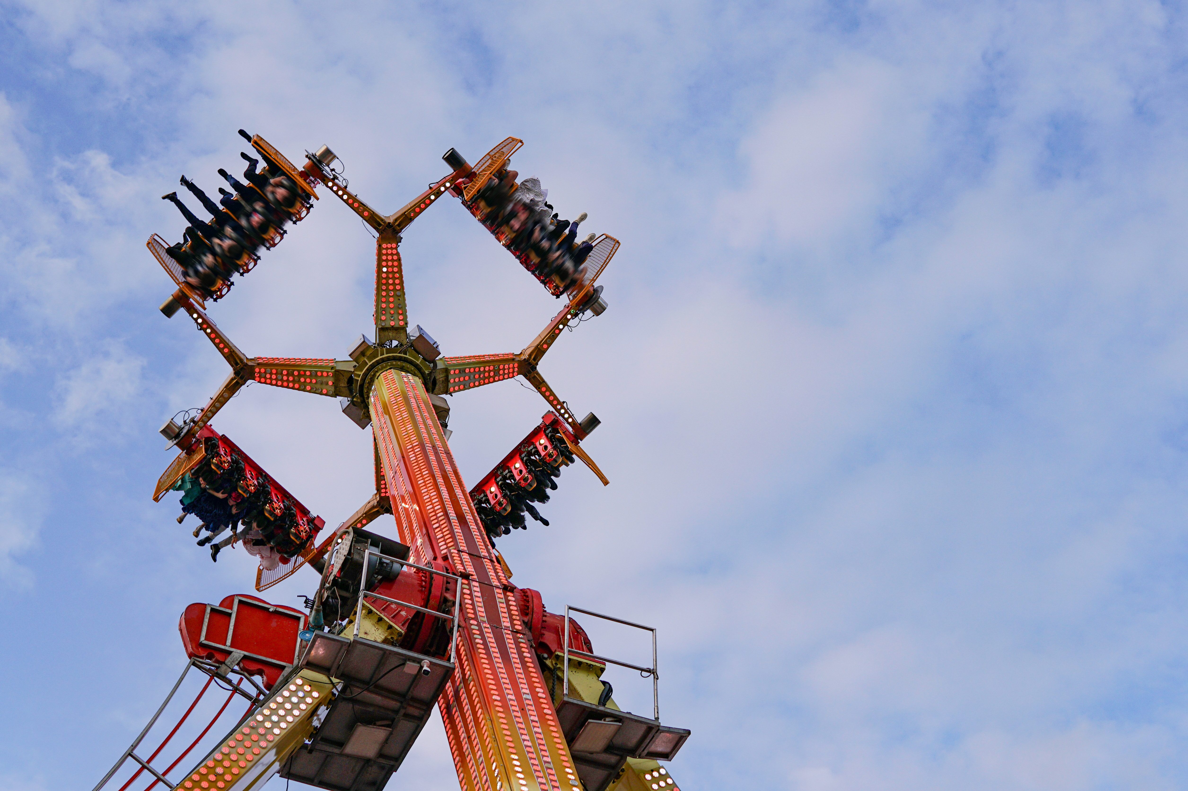A view looking up at a tall amusement park ride, featuring a central pole with multiple arms extending outward. Each arm has seats with passengers suspended upside down, and the structure is adorned with bright lights.