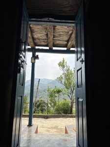 A view through an open blue door, revealing a rural landscape with green trees and mountains in the background. 