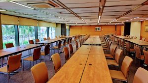 A spacious conference room featuring multiple long tables arranged in rows, accompanied by chairs. Large windows along one side allow natural light to illuminate the space, and palm trees can be seen outside. 