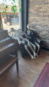 Two plastic skeletons reclining in a chair in front of a window and beside a piano and a brick wall in a coffee shop.
