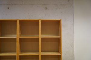 A wooden bookshelf with empty shelves, set against a textured concrete wall.