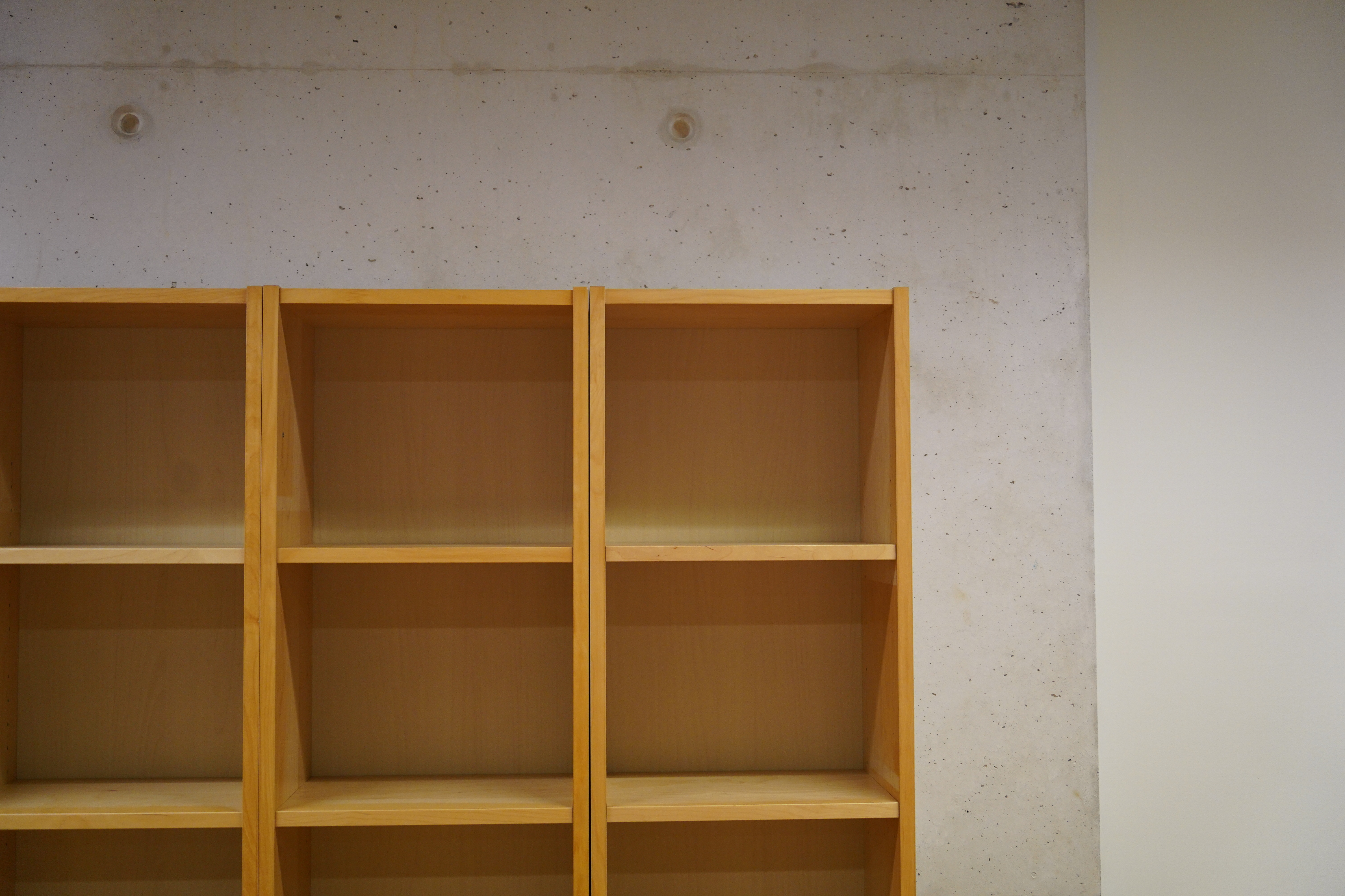 A wooden bookshelf with empty shelves, set against a textured concrete wall.