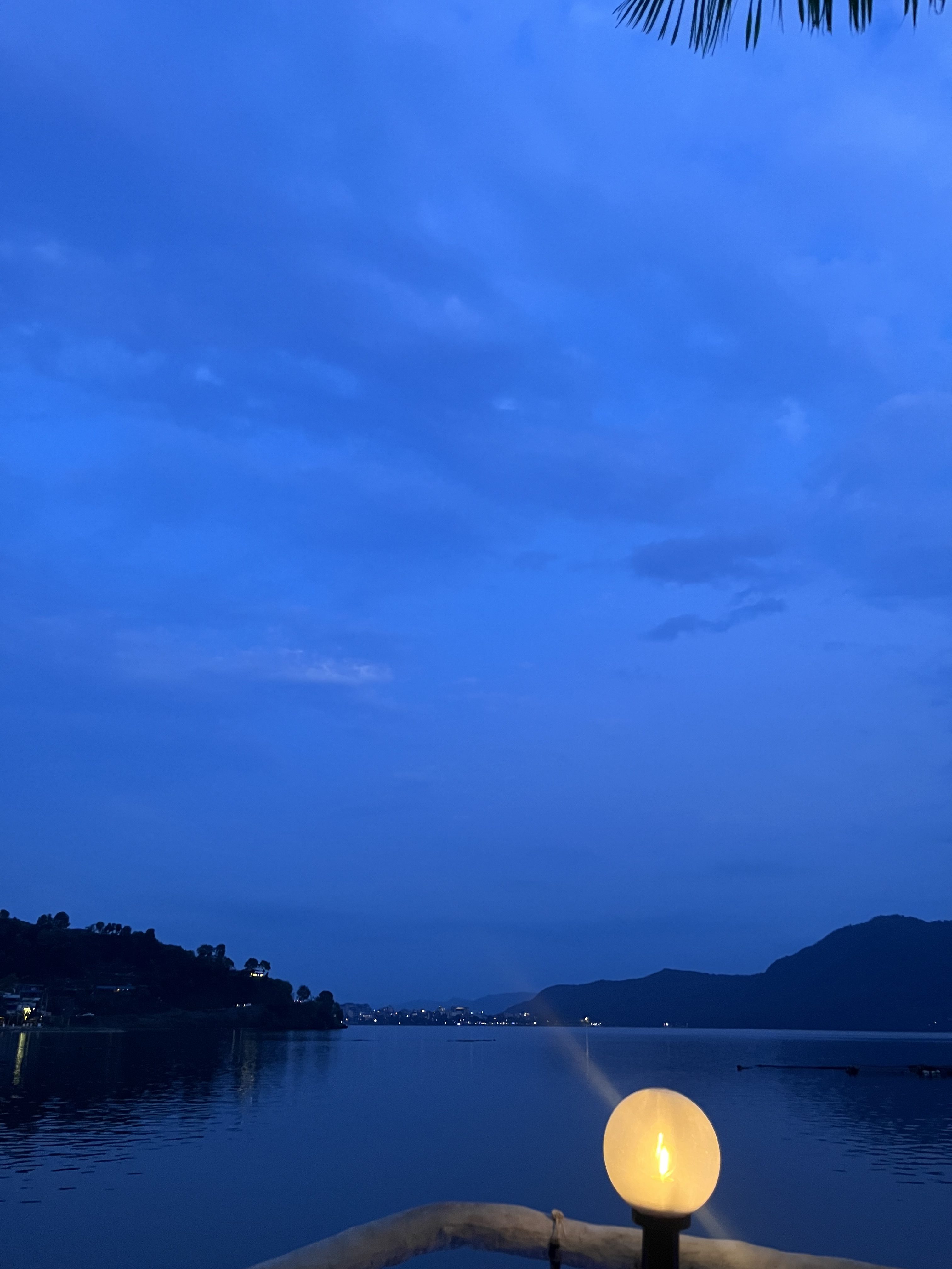 A serene twilight scene by a calm lake, with a softly glowing round lamp in the foreground. The sky is a gradient of deep blue hues, with scattered clouds