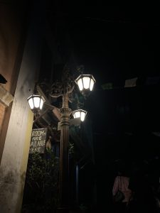 Three street lamps on one wrought iron lamppost. The scene is dark, and the lights illuminate a sign that has "Rooms are available here" scrawled on it.