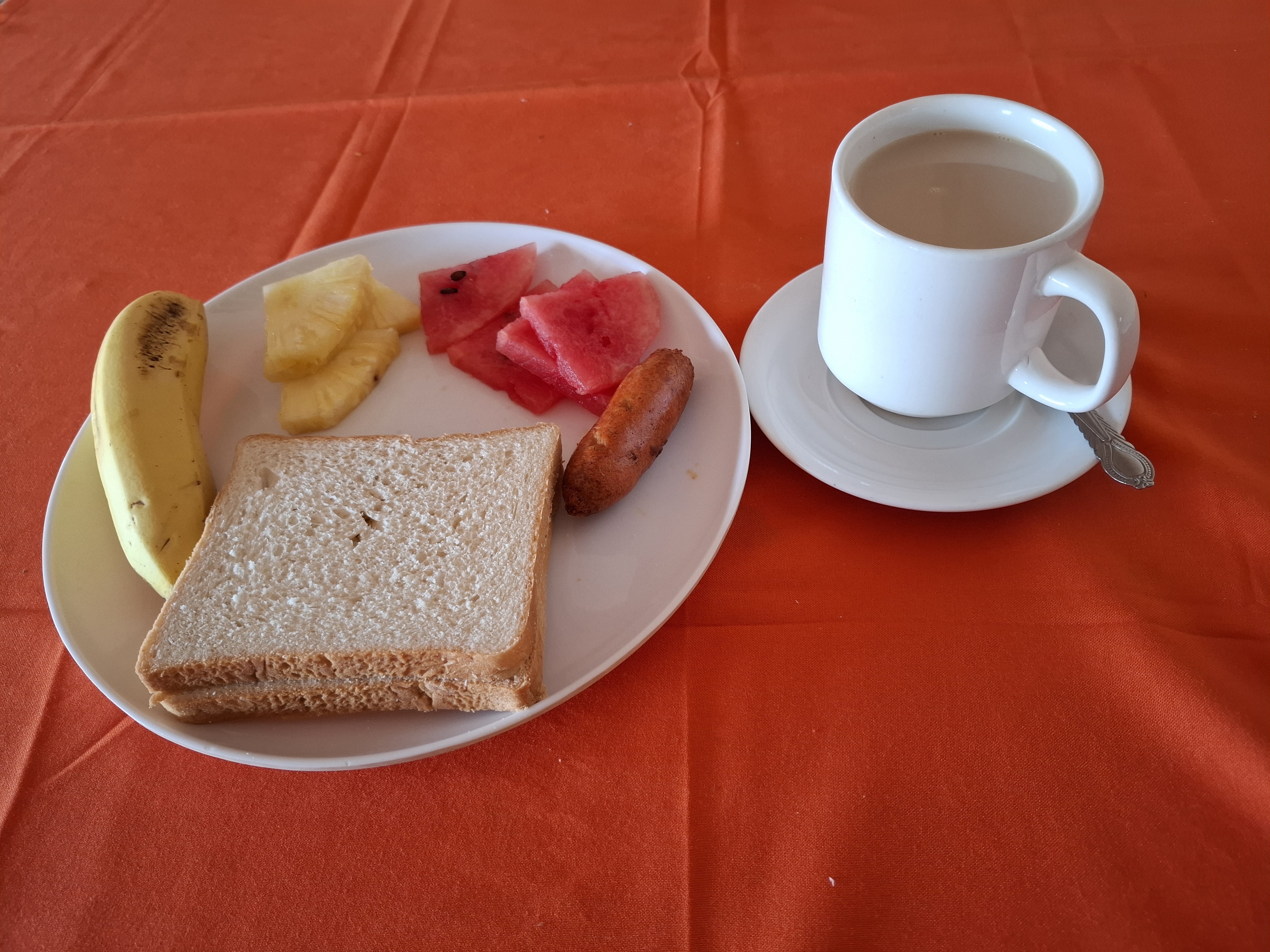 A plate with a variety of breakfast items including a banana, pieces of pineapple, watermelon slices, a slice of bread, and a small sausage.