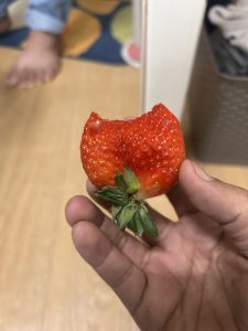 A close-up image of a partially eaten strawberry held in a hand. The bright red fruit shows a bite mark on one side, and the green leaves remain attached at the base. The background is softly blurred, revealing a wooden surface.