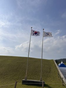 Two flags are flying on poles in front of a grassy hill. The flag on the left is the national flag of South Korea, featuring a red and blue yin-yang symbol in the center surrounded by black trigrams. 