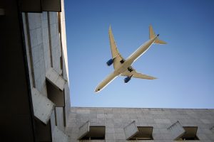 An airplane is flying overhead against a clear blue sky, captured from below between the walls of a modern building that features a textured concrete facade.