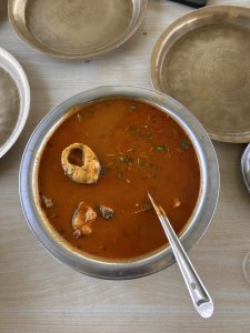 A large, circular steel bowl contains a rich, reddish-brown soup with pieces of fish and herbs floating on the surface.