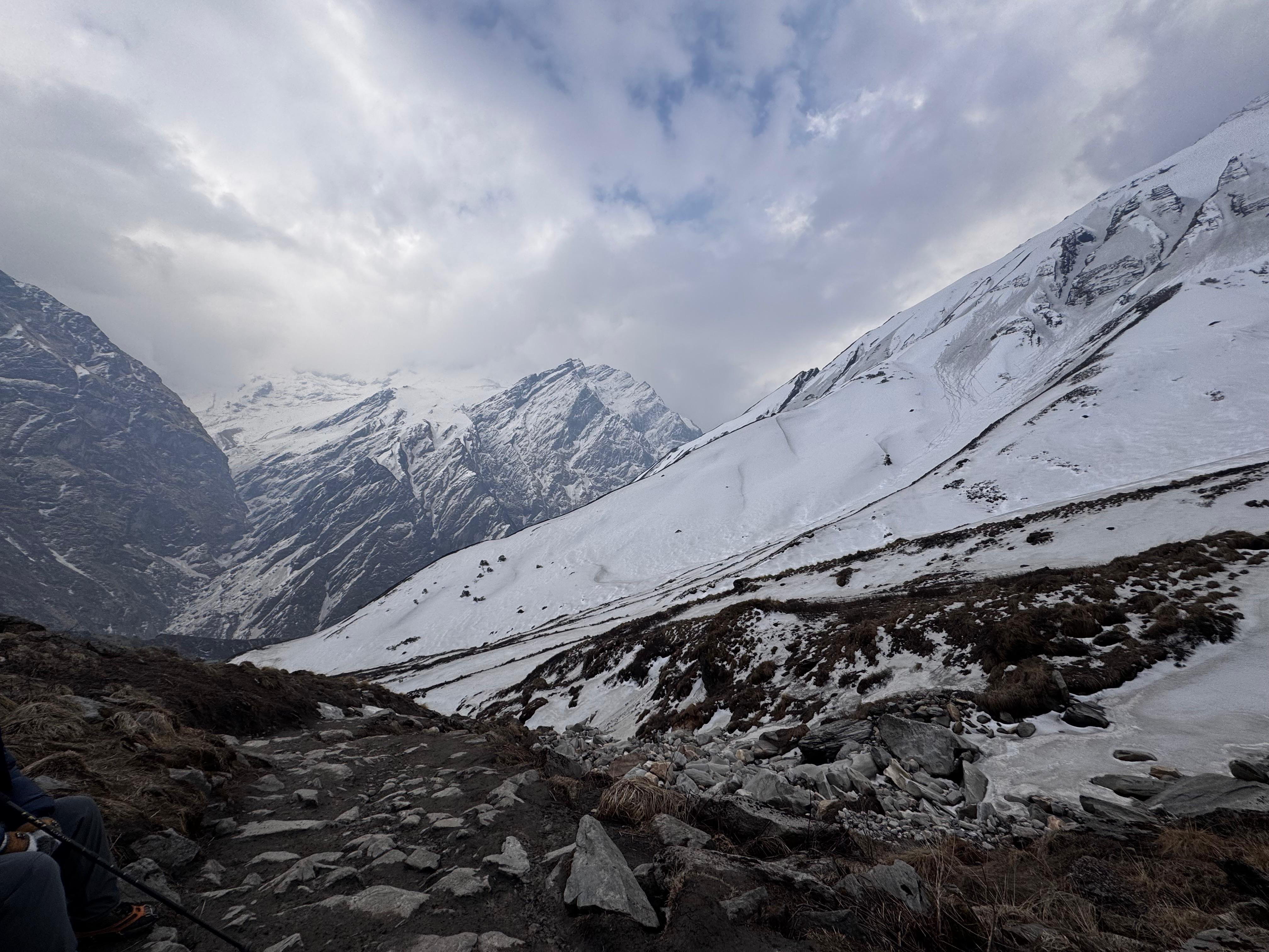 A scenic view of snow-covered mountains under a cloudy sky. Jagged peaks rise prominently in the distance, while the foreground features rocky terrain and patches of snow.