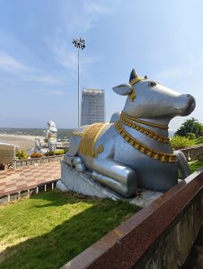 The image shows a large, silver-colored statue of Nandi, the sacred bull associated with the Hindu god Shiva, at the Nandi Temple in Murudeshwar, Karnataka.
