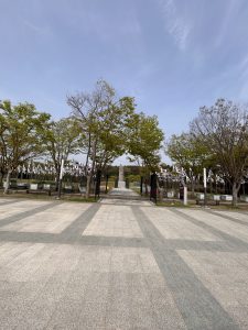 A wide view of a park entrance featuring a pathway made of square tiles leading towards a monument in the distance. Flanking the pathway are trees, some with green leaves and others with bare branches.