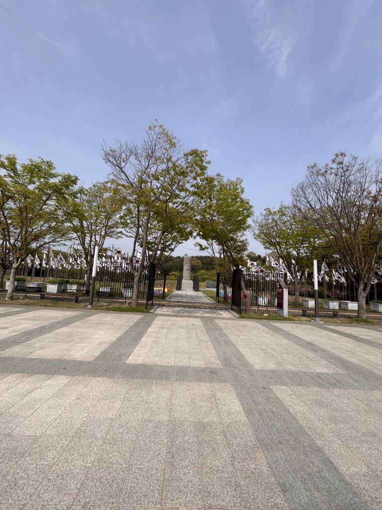 A wide view of a park entrance featuring a pathway made of square tiles leading towards a monument in the distance. Flanking the pathway are trees, some with green leaves and others with bare branches.