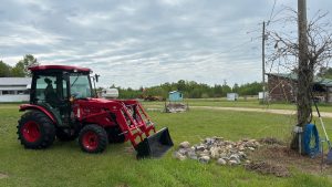 A farmyard scene with a large red tractor dominating the left side of the image.  It has a bucket on the front and on the ground in front of it are a bunch of large cobbles.