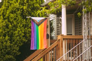 A slightly frayed Progress Pride flag hangs partially rolled from a horizontal pole in front of a house with wooden stairs. A large tree stands in the background, framing the scene.
