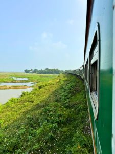 A view from a train window, showcasing lush green vegetation and patches of water alongside the railway track. 
