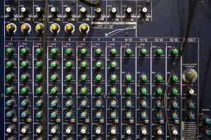 A close-up view of a professional audio mixer featuring numerous knobs and buttons. The controls include gain, compression, and equalization settings, with various labeled channels.