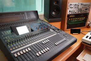 A close-up view of an audio mixing console featuring a variety of faders, knobs, and buttons. The console includes a digital display and several illuminated indicators. To the right, there is a rack of audio equipment with multiple modules, and behind it, a black studio monitor speaker stands on a wooden surface.