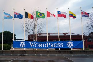 A row of nine flags representing different countries, including Argentina, Australia, Belgium, Brazil, Canada, Chile, Columbia, Costa Rica, are displayed on tall poles against a backdrop of a building with solar panels on the roof. A large blue banner with the word "WordPress" is positioned below the flags. 