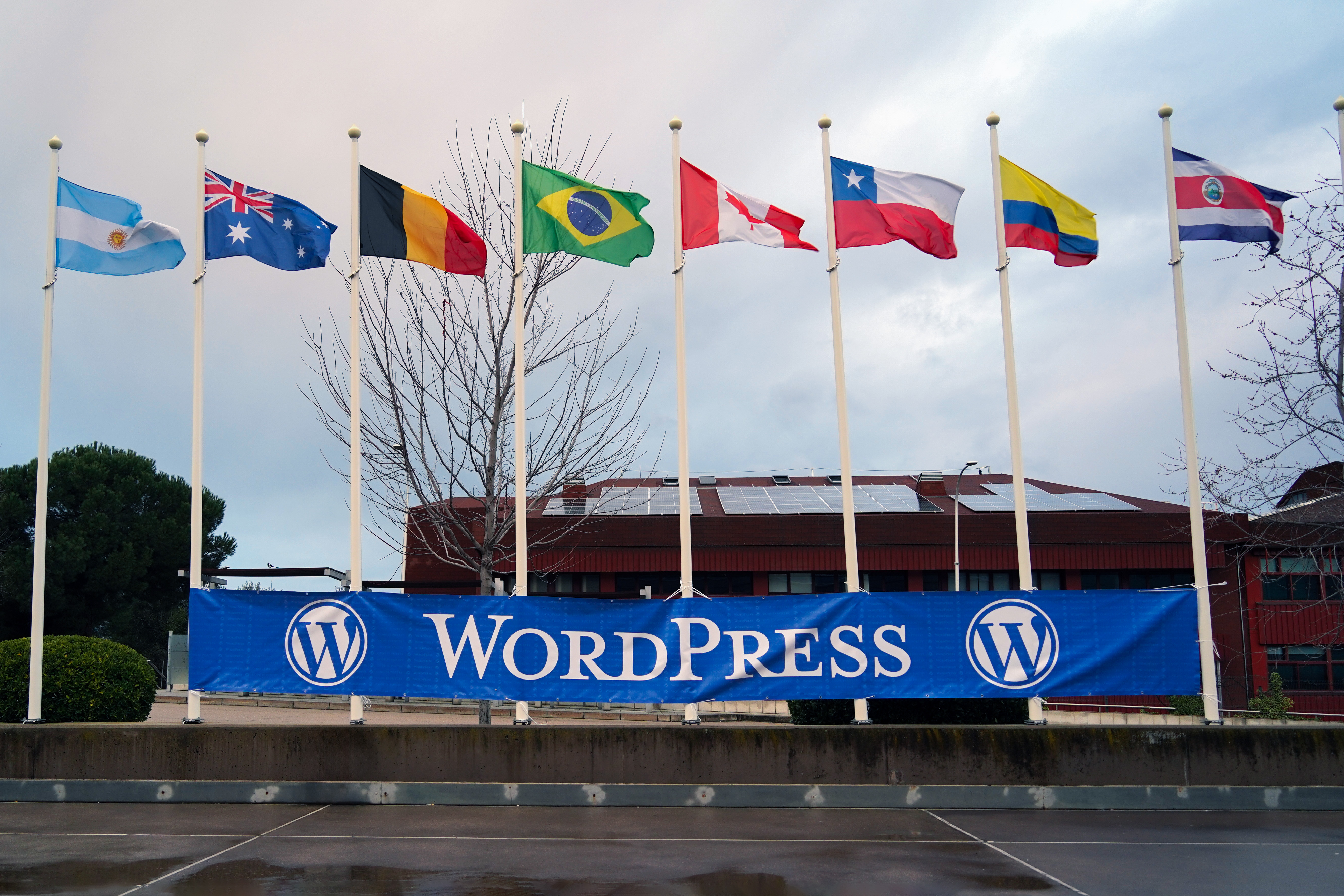 A row of nine flags representing different countries, including Argentina, Australia, Belgium, Brazil, Canada, Chile, Columbia, Costa Rica, are displayed on tall poles against a backdrop of a building with solar panels on the roof. A large blue banner with the word "WordPress" is positioned below the flags.
