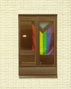 A Progress Pride flag hangs in a window of cream brick building.