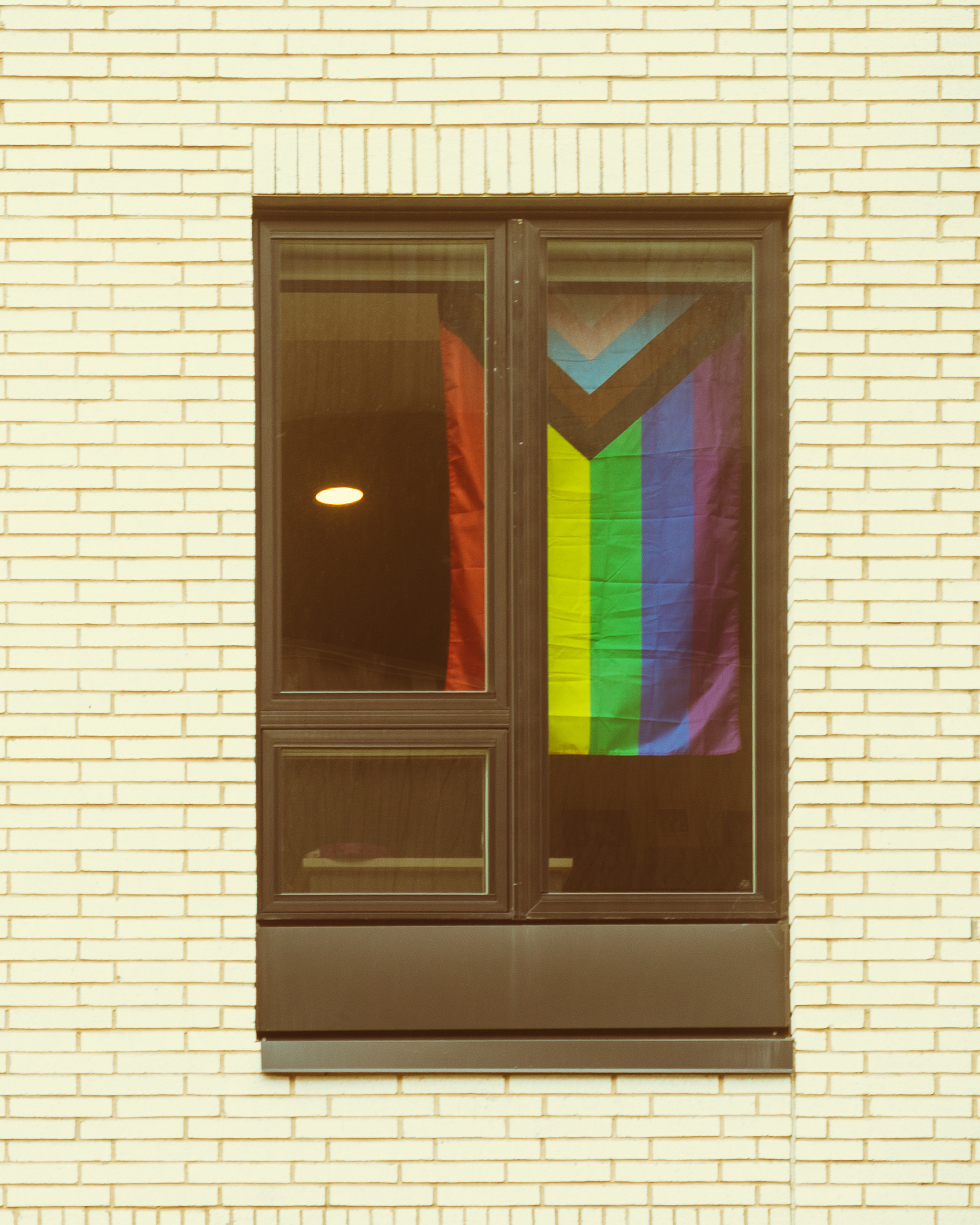 A Progress Pride flag hangs in a window of cream brick building.