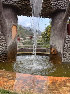 A decorative water fountain designed to resemble a tree structure. Water cascades from a spout at the top and flows into a circular basin that has a slight green tint.
