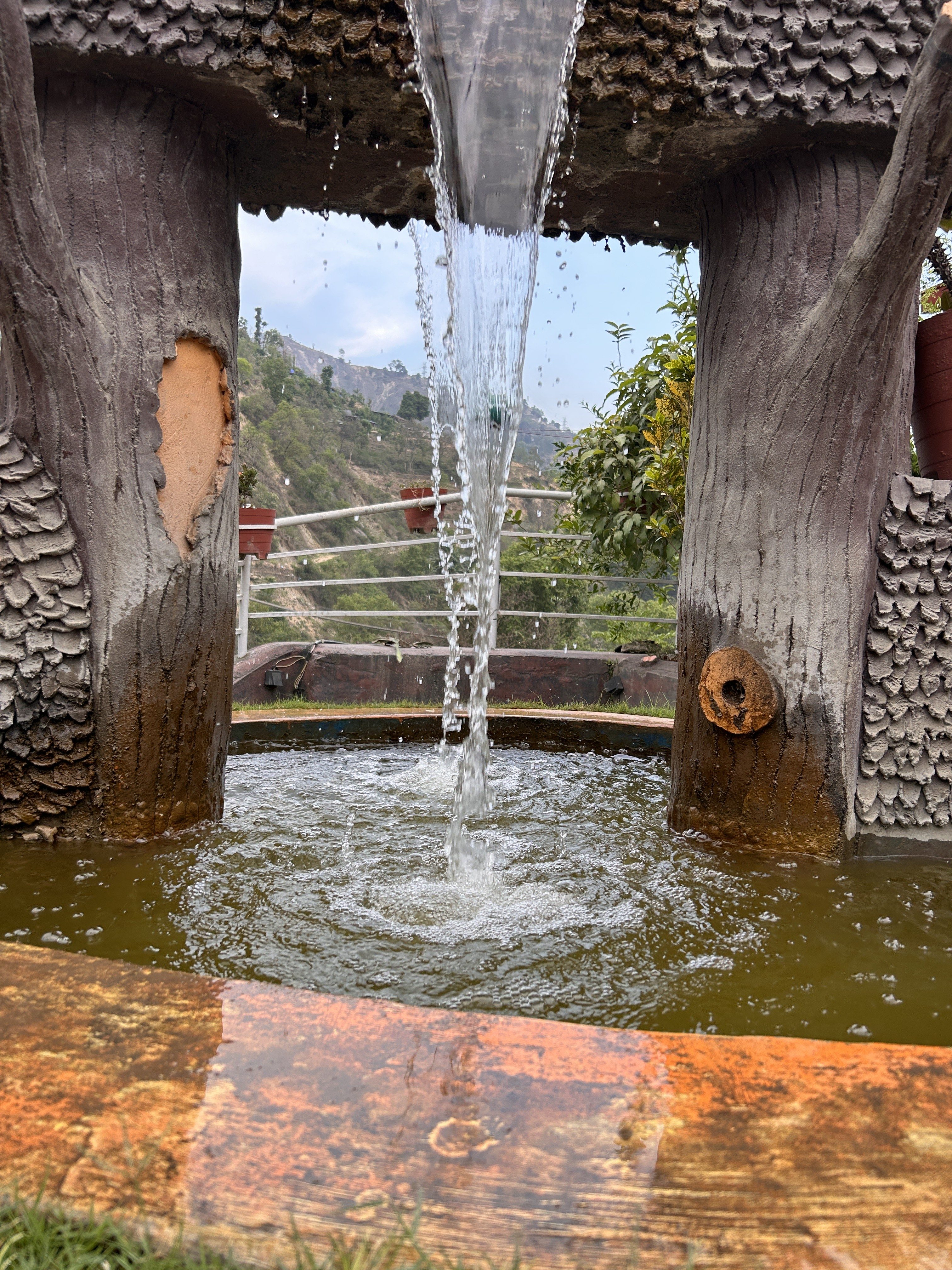 A decorative water fountain designed to resemble a tree structure. Water cascades from a spout at the top and flows into a circular basin that has a slight green tint.